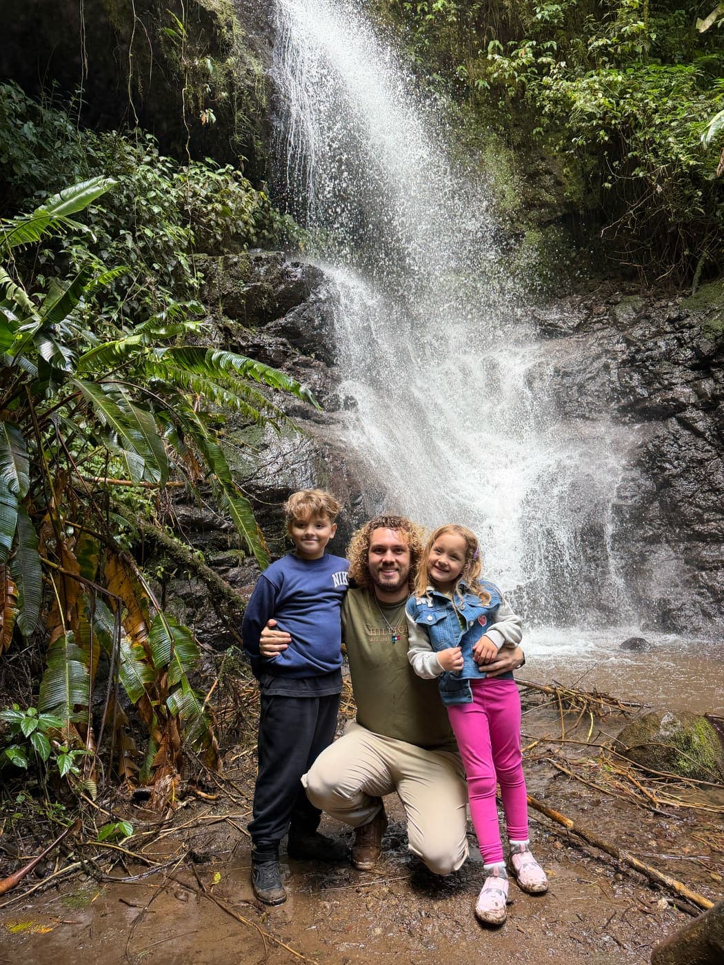 Jose crouched between his two children in front of a rainforest waterfall