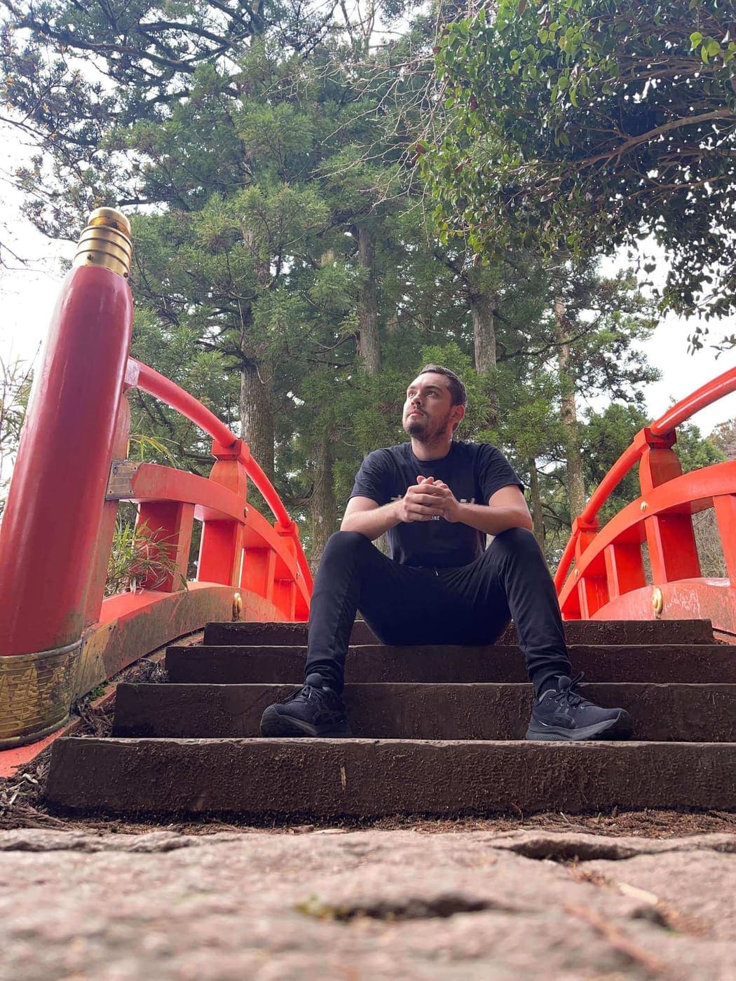 Jose sitting on stone steps beside a red bridge in a Japanese cedar forest